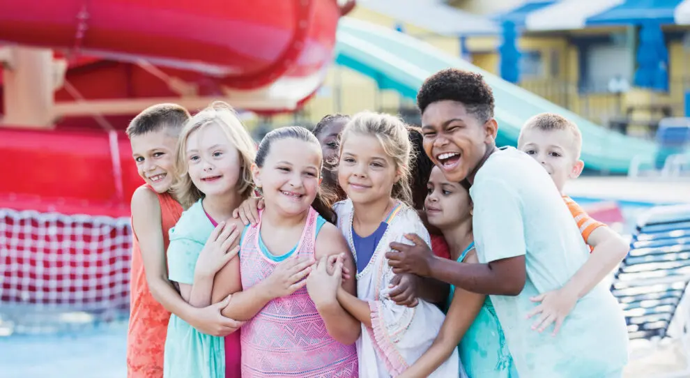 A multi-ethnic group of children, 8 to 11 years old, having fun together at a water park. They are hugging one another, smiling at the camera. The girl in light green, second child from the left, has down syndrome.